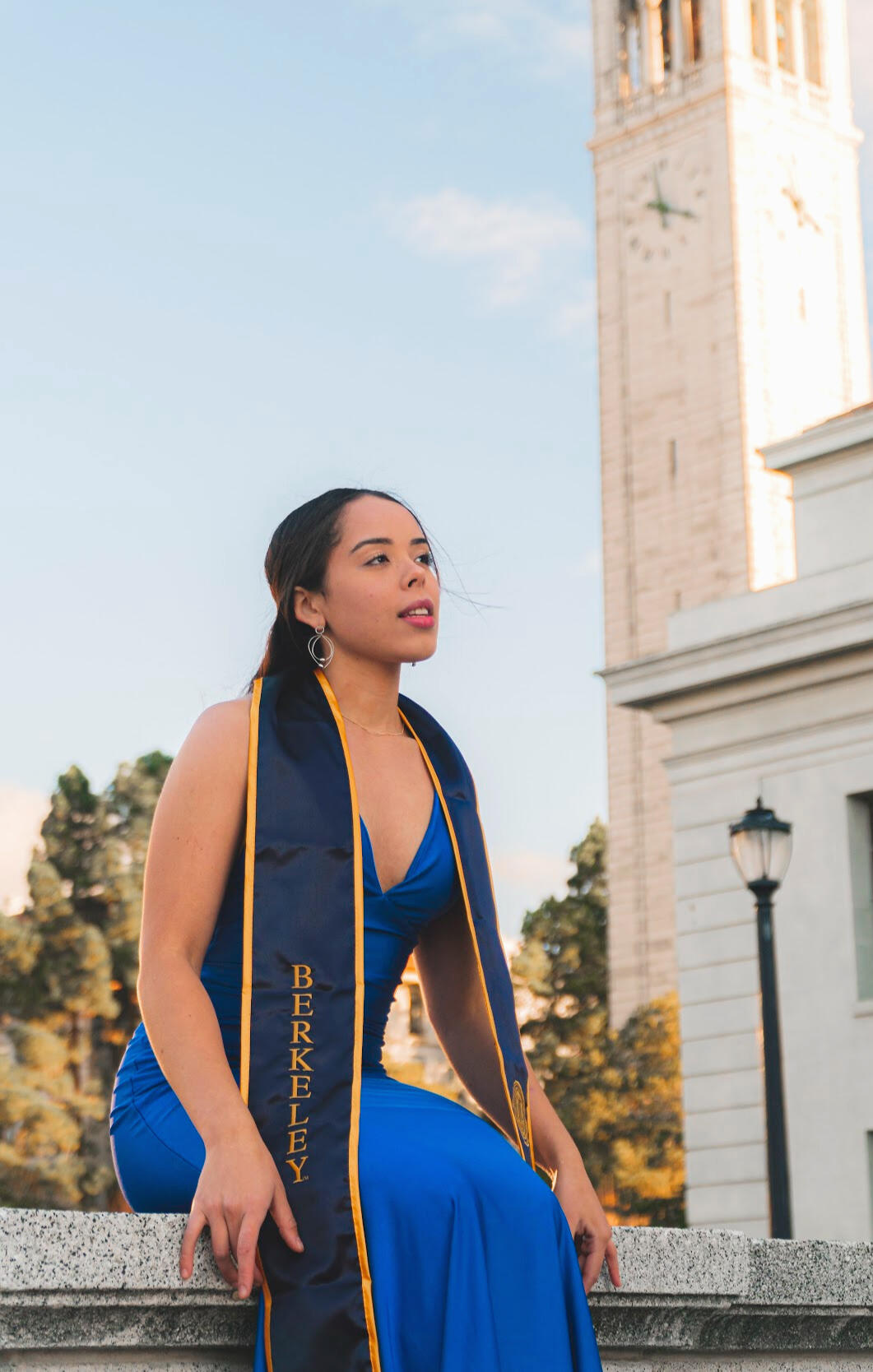 An excited young girl in a vibrant blue dress, adorned with UC Berkeley stoles, sitting on a bench with the iconic UC Berkeley clock tower in the background