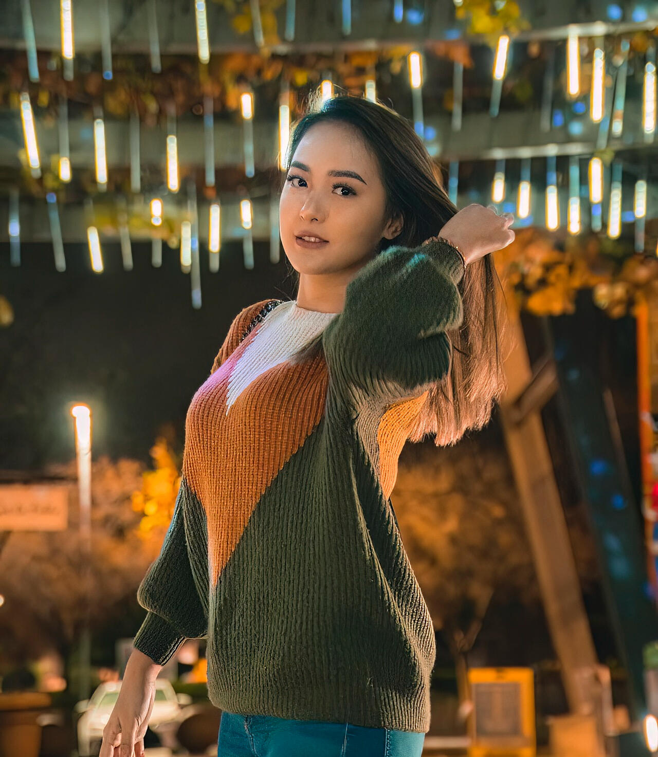 An enchanting evening portrait of a young woman in a cozy woven sweater, gazing into the camera amidst the shimmering backdrop of decorative lights