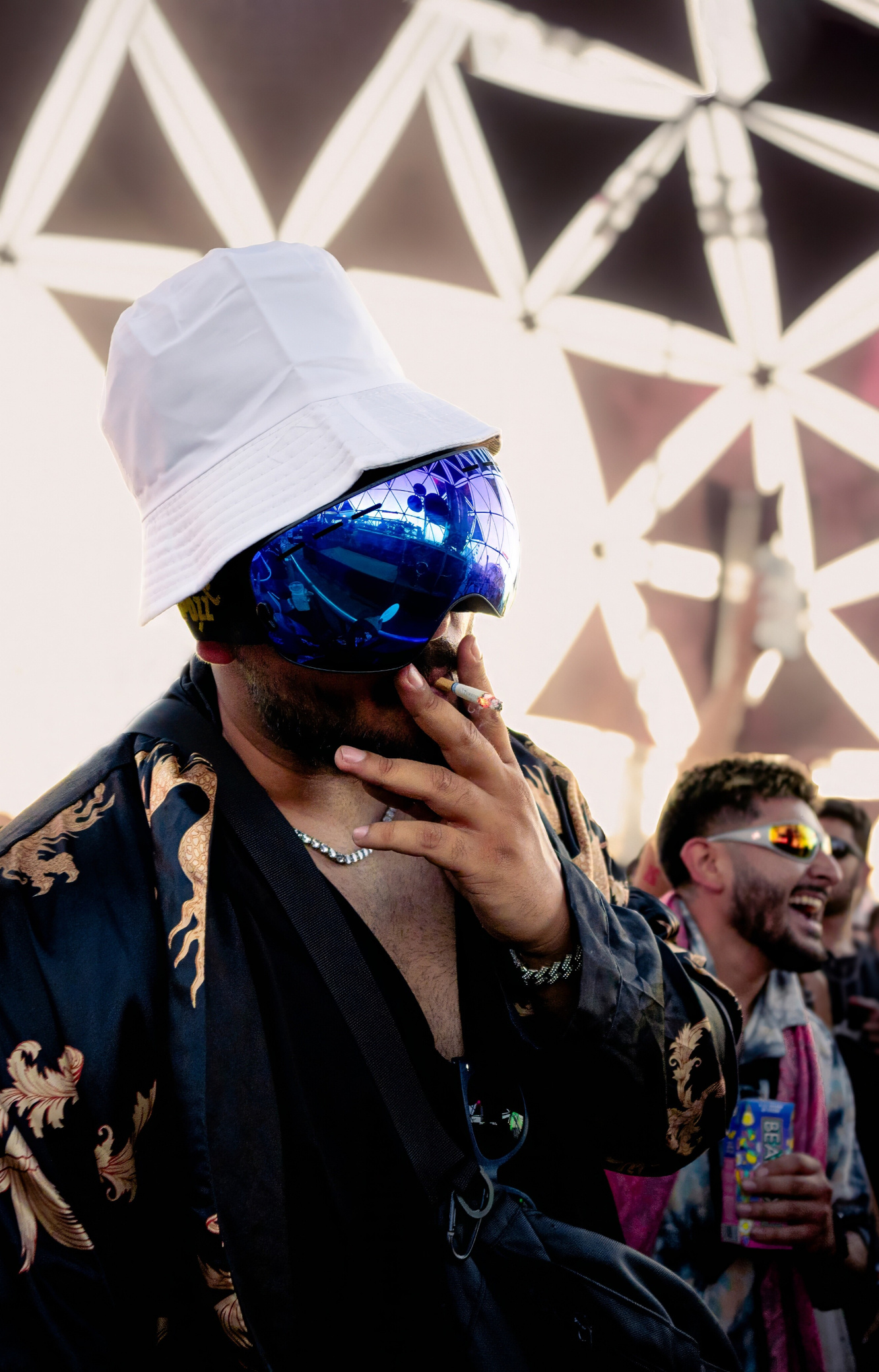 Man wearing purple ski goggles and a white bucket hat smoking a cigarette at an EDM festival.