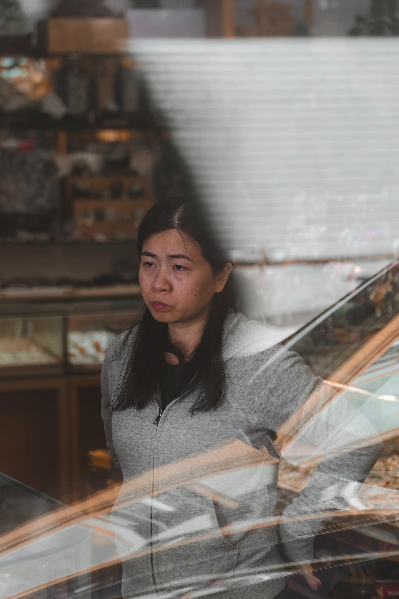 An image of an Asian female shop owner looking out the window of her jewelry store with reflections of the street visible